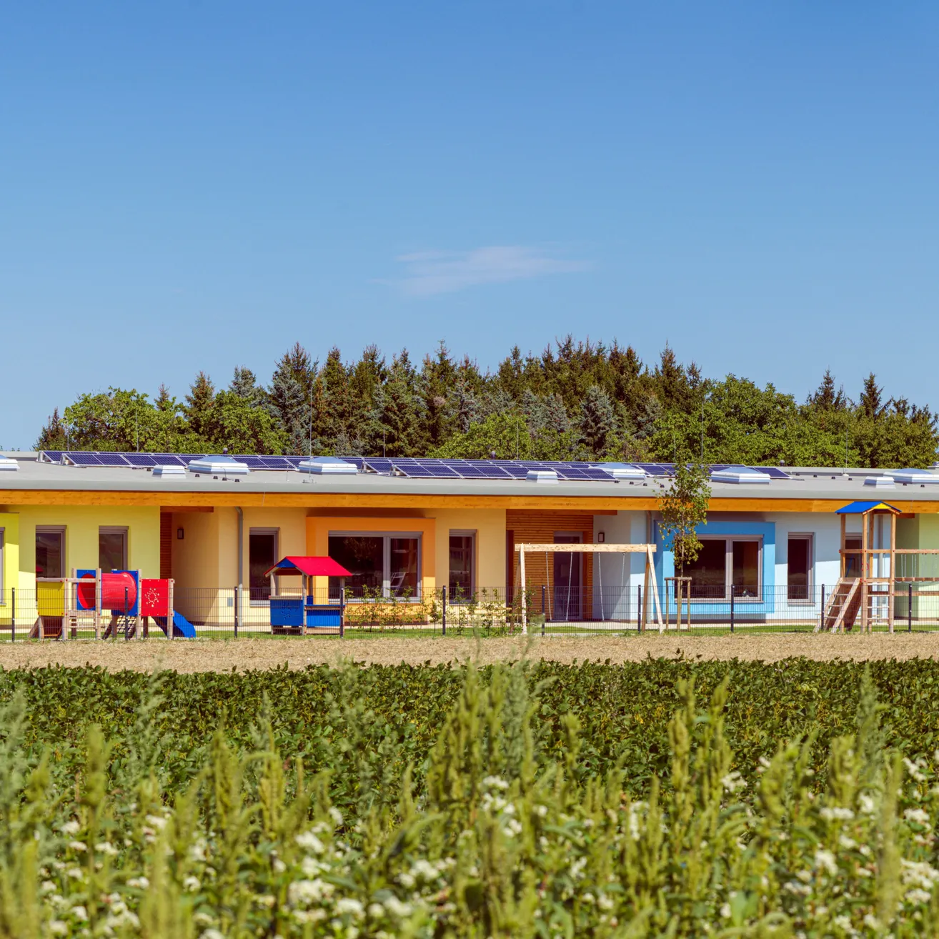 Ein modernes, gelbes Gebäude mit Solarpanelen auf dem Dach und einem Spielplatz davor.