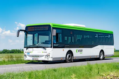 Ein weißer und grüner Bus mit dem Wort 'Vor' auf der Seite fährt auf einer Straße mit einem Grasfeld auf der Seite.
