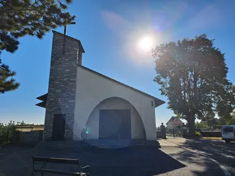 Eine kleine Kapelle mit einem Steinturm und einem Kreuz auf der Spitze, unter einem hellen Sonnenschein.