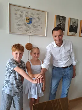 Ein Mann und zwei Kinder stehen vor einer Wand mit einem gerahmten Dokument und drei gerahmten Porträts.