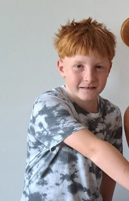 Ein junger Junge mit roten Haaren und Sommersprossen steht gegen eine weiße Wand und trägt ein Tie-Dye-T-Shirt.