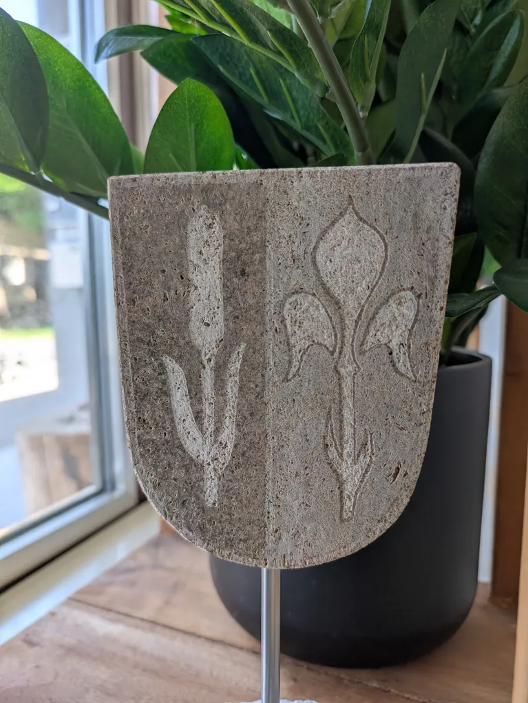 Abschnittsbild: Eine Betonplatte mit Fleur-de-Lis-Design vor einer grünen Topfpflanze in der Nähe eines Fensters.
