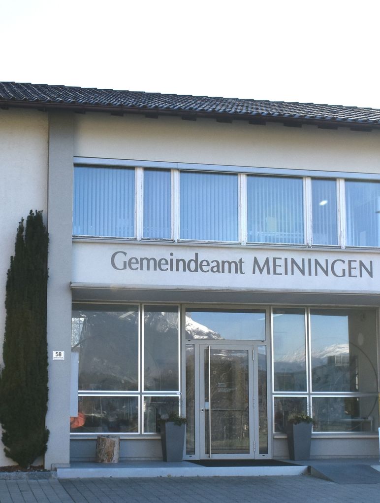  Abschnitt Bild: Das Gemeindeamt Meiningen Gebäude ist weiß mit einem schwarzen Dach. Es hat Glasfenster, eine Glastür und ein Schild mit der Aufschrift 'Gemeindeamt Meiningen'. Es gibt Topfpflanzen und die Nummer 58 auf der linken Seite.
