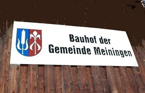 Ein weißes Schild, das an einem Holzzaun befestigt ist, zeigt den Text 'Bauhof der Gemeinde Meiningen'. Das Schild hat ein blaues und rotes Emblem mit einem weißen blumenartigen Design auf der rechten Seite.