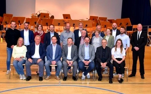 Eine Gruppe von Männern und Frauen in formeller Kleidung posiert für ein Foto auf einer Bühne mit Musikinstrumenten im Hintergrund.