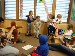 Mehrere Kinder in einem Raum, eine Person hält einen Affenplüsch und einige Erwachsene sitzen auf dem Boden.