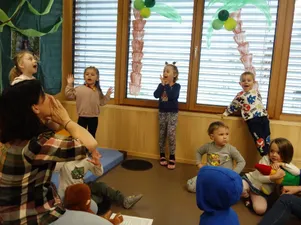 Eine Gruppe junger Kinder ist in einem Raum mit einer Holzwand und großen Fenstern zu sehen. Eines der Kinder tanzt, während die anderen sitzen und zusehen. Eine Frau steht vor den Kindern und klatscht.