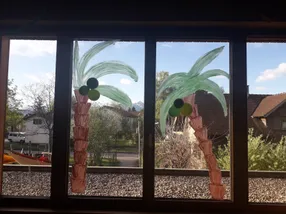 Zwei Fenster, die mit grünen Palmen aus Luftballons verziert sind. Ein Fenster hat grüne und schwarze Ballons, das andere grüne und gelbe Ballons.