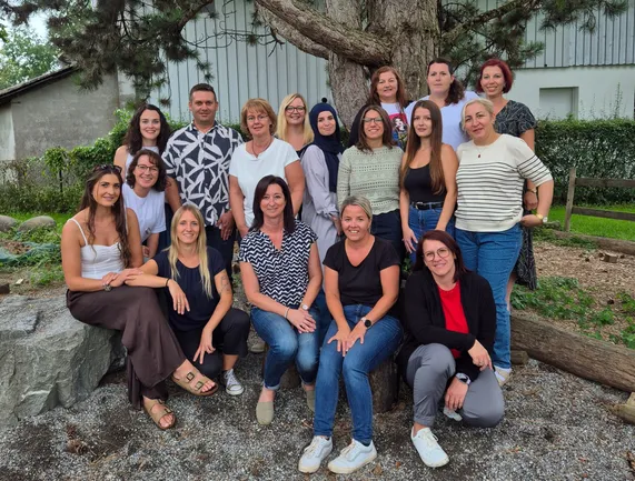 Eine vielfältige Gruppe von Menschen, darunter Frauen und Männer, posiert für ein Foto in einem Park mit einem Baum im Hintergrund.