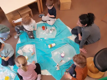 Kinder sitzen um ein blaues Tuch auf dem Boden und spielen mit Spielzeug und Bechern mit Wasser. Eine Frau beobachtet sie von der Seite.