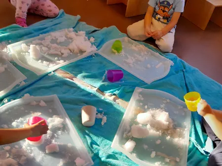 Ein Kind sitzt auf einem blauen Handtuch und spielt mit Schnee in Plastikwannen, mit Becher und Wasser in der Nähe.