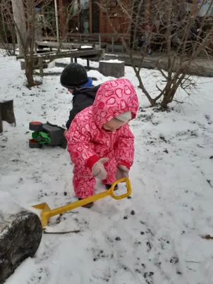 Zwei Kinder in Schneeanzügen spielen im Schnee. Ein Kind benutzt einen gelben Schneeschaufel. Hinter ihnen stehen Bäume, eine Holzbank und ein Baumstamm.