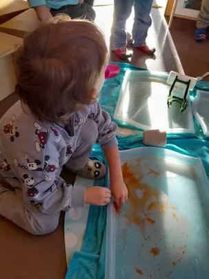 Ein Kind spielt mit einer Wasserwanne mit orangefarbener Flüssigkeit, trägt Pyjamas mit Mickey-Mouse-Designs, während eine Person in roten Schuhen in der Nähe steht.
