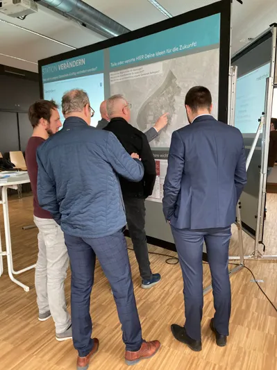 Fünf Männer in Geschäftskleidung stehen um eine Projektionsfläche herum und diskutieren zukünftige Ideen. Eine große Karte ist auf dem Bildschirm mit einer Textüberlagerung angezeigt.