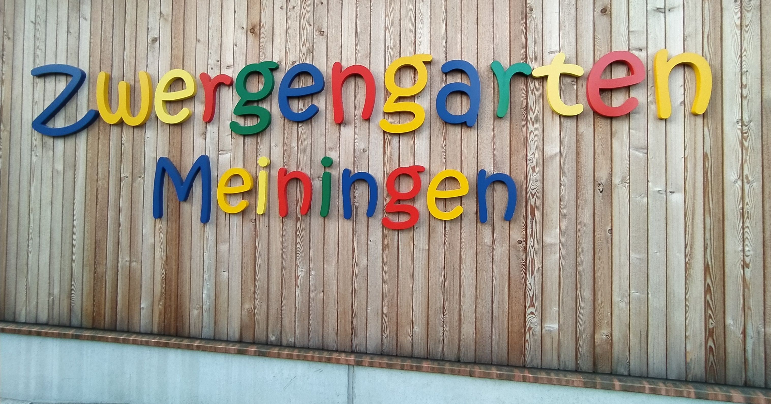Ein Holzschild mit dem Wort 'gergengart' in farbenfrohen Buchstaben auf einer Holzwand. Darunter steht das Wort 'Meiningen' in Regenbogenfarben.