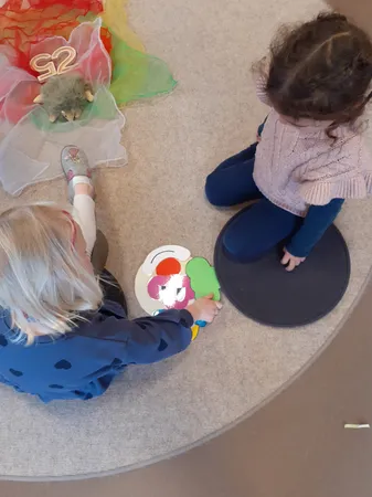 Zwei Kinder spielen auf dem Teppichboden. Ein Kind hält ein farbenfrohes Eistüten-förmiges Puzzleteil. Ein anderes Kind sitzt in der Nähe. Es gibt Spielzeug und bunte Tüll auf dem Boden.