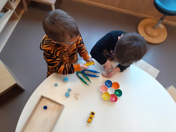 Zwei kleine Jungen sitzen an einem Tisch und beschäftigen sich mit bunten Gegenständen. Ein Junge trägt ein Tiger-gestreiftes Shirt, der andere ein schwarzes. Der Tisch ist mit verschiedenen Spielzeugen und Bastelmaterialien übersät.