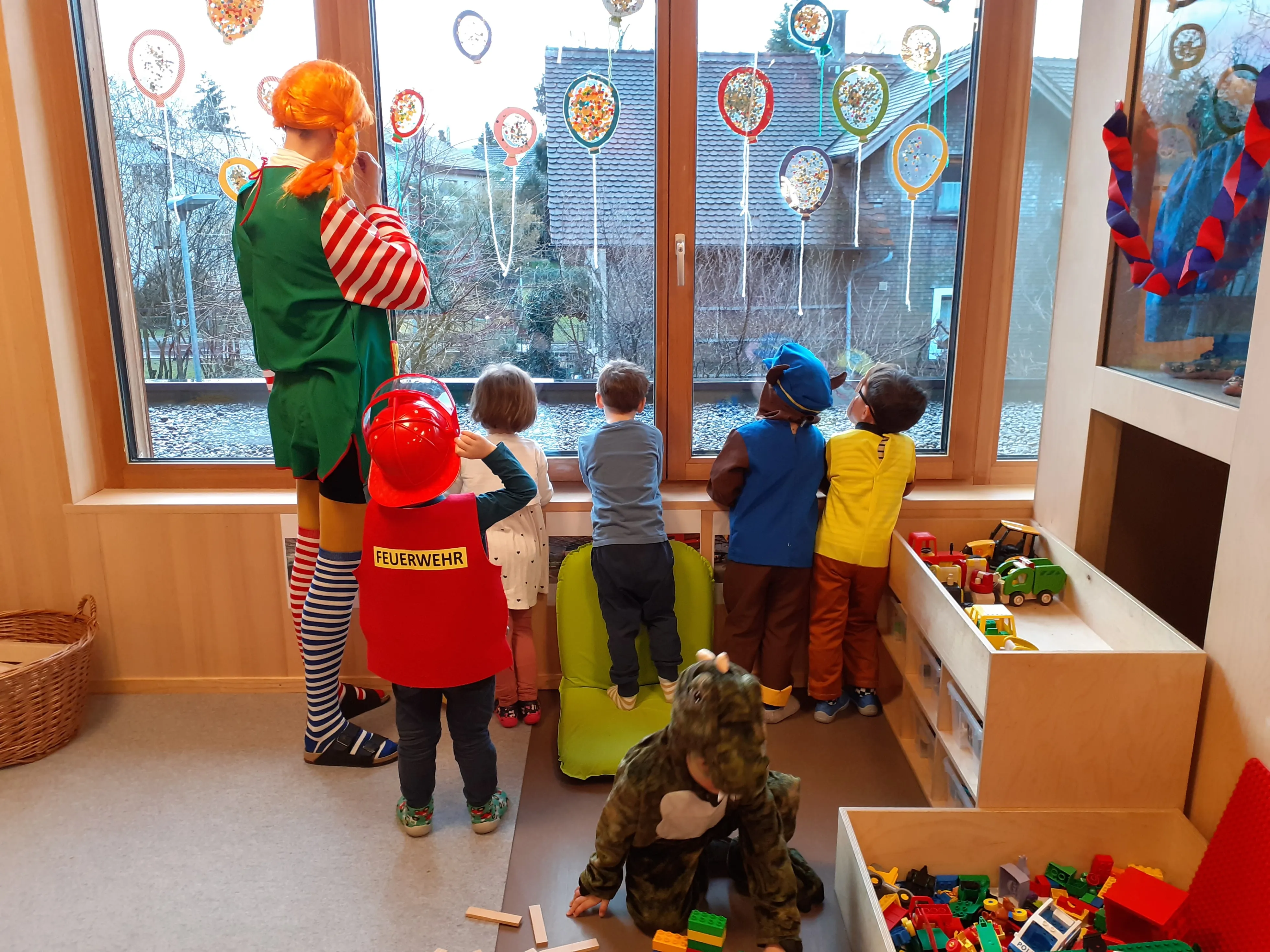 Eine Gruppe von Kindern in Kostümen schaut aus einem Fenster mit bunten Luftballons. Eine Frau in einer Clown-Kostümierung steht hinter ihnen.