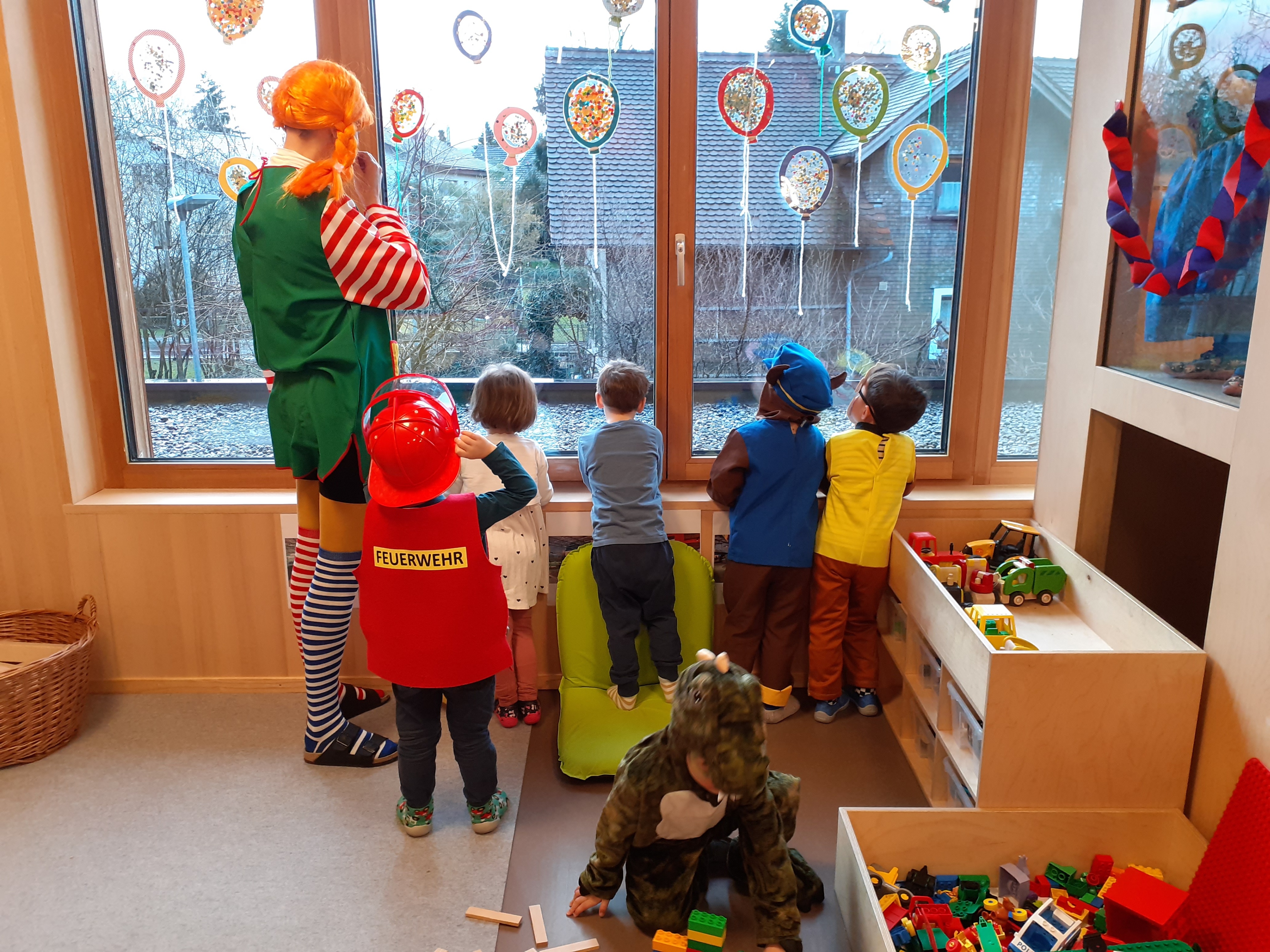 Eine Gruppe von Kindern in Kostümen schaut aus einem Fenster mit bunten Luftballons. Eine Frau in einer Clown-Kostümierung steht hinter ihnen.