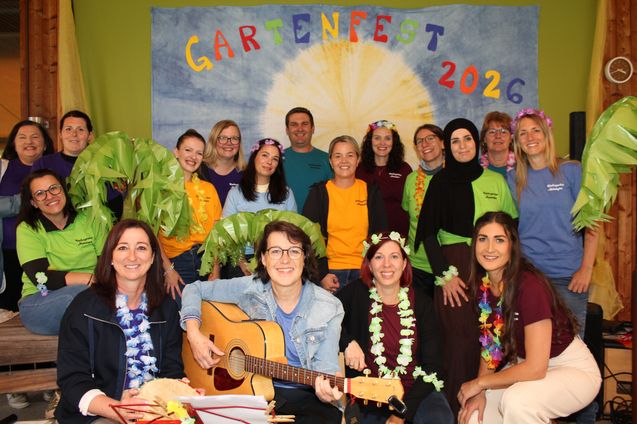 Eine Gruppe von Frauen und Männern posiert für ein Foto vor einem Banner, auf dem Gartenfest 2026 steht. Sie tragen alle gelbe und grüne Hemden. Einige halten Gitarren und andere tragen Blumenkränze. Sie alle lächeln.