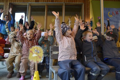 Eine Gruppe junger Kinder hebt die Hände und lächelt, einige tragen Mützen. Sie scheinen in einem Klassenzimmer mit Glaswänden zu spielen.