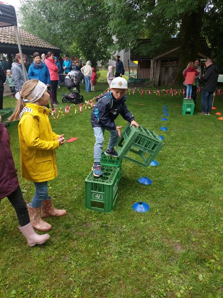 Kinder nehmen an einem Hindernisparcours auf einem Rasenfeld mit grünen Kisten teil. Zuschauer und ein Kinderwagen sind im Hintergrund.
