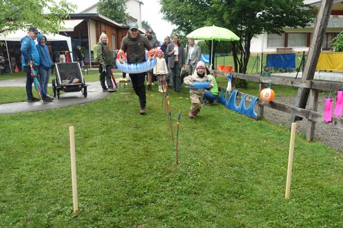 Eine Gruppe von Menschen nimmt an einer Veranstaltung im Freien teil, wobei ein Mann läuft, während er eine aufblasbare Röhre trägt, und ein Kind vor ihm steht. Einige andere stehen in der Nähe, einige beobachten und andere beschäftigen sich mit Aktivitäten.