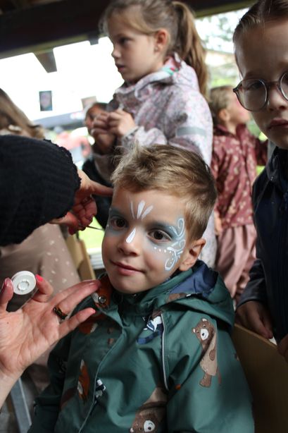 Ein Kind lässt sich in einem festlichen Umfeld das Gesicht bemalen, während mehrere Kinder zuschauen.