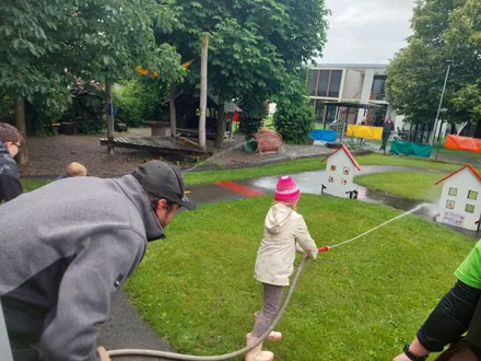 Ein Mann und ein Kind spielen mit einem Schlauch in einem Park mit einem hölzernen Spielhaus und einem Gebäude im Hintergrund.