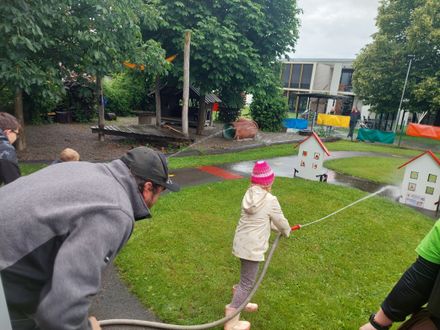 Ein Mann und ein Kind spielen mit einem Schlauch in einem Park mit einem hölzernen Spielhaus und einem Gebäude im Hintergrund.