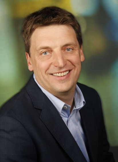 Ein lächelnder Mann in einem dunklen Anzug und hellblauem Hemd, posiert für ein professionelles Foto mit einem unscharfen grünen Hintergrund.