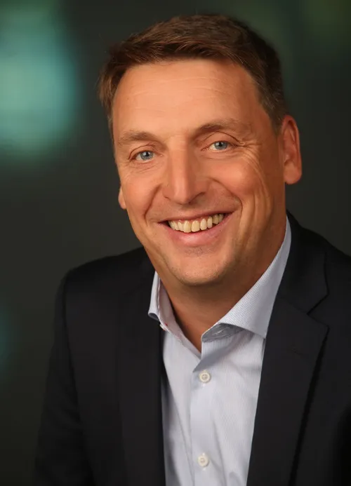 Ein lächelnder Mann in einem Anzug mit hellblauem Hemd unter einem dunklen Mantel, Zähne zeigend, mit einem unscharfen Hintergrund.