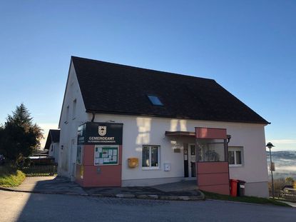 Die Vorderansicht eines weißen Gebäudes mit schwarzem Dach. Das Gebäude hat einen Aushang mit angehefteten Papieren. Es gibt zwei Mülleimer und eine Glastür. Der Himmel ist klar und blau.