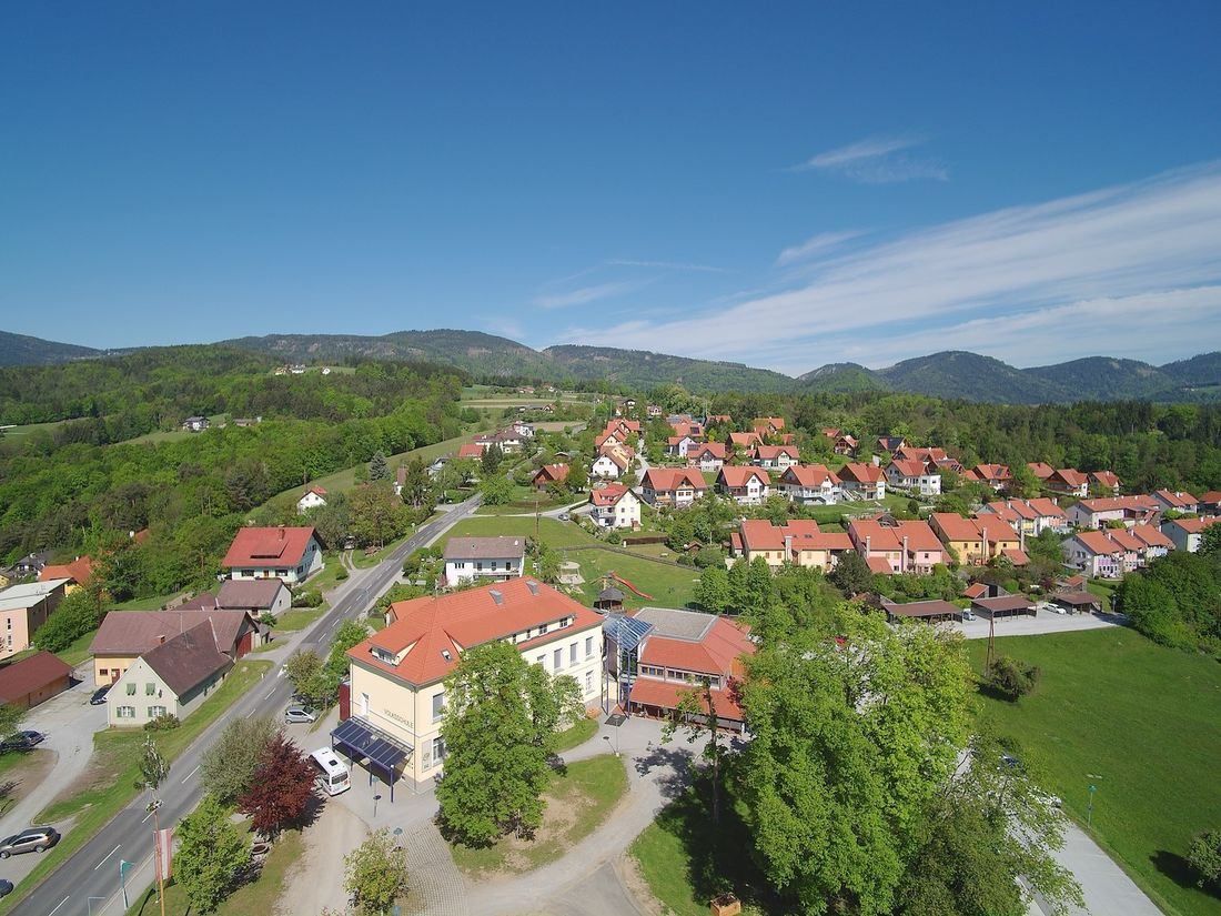 Luftaufnahme eines Dorfes mit zahlreichen Häusern und Gebäuden, umgeben von viel Grün und Bergen unter einem klaren blauen Himmel.