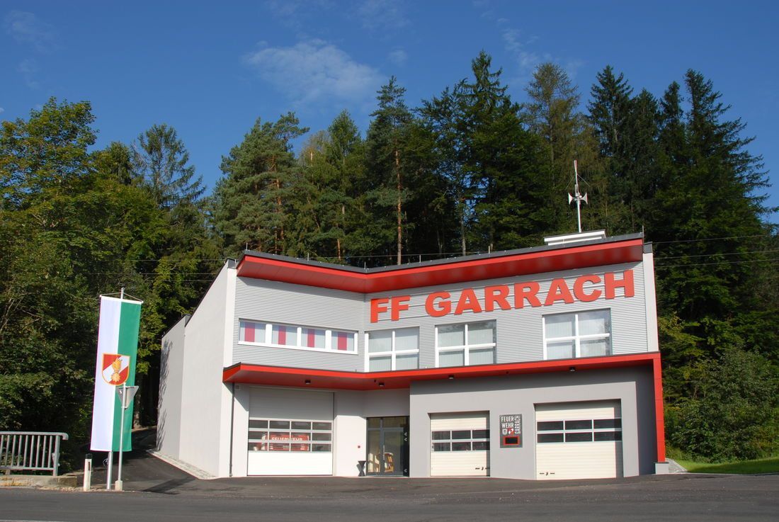Ein weisses Gebäude mit rotem Rand heißt FF Garrach. Es hat drei Garagen, Glastüren und Fenster. Hohe Bäume umgeben das Gebäude und der Himmel ist klar und blau.