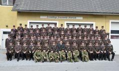 Eine Gruppe von Militärangehörigen in Uniform, die vor einem gelben Gebäude mit zwei Garagen stehen. Sie posieren für ein Foto, wobei mehrere Personen in der vorderen Reihe sitzen.