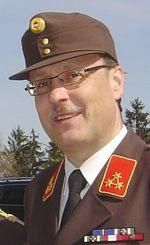 Ein Mann in einem Militäruniform mit einem braunen Hut, Brille, einem roten Abzeichen und mehreren Medaillen steht draußen. Der Hintergrund ist unscharf mit Bäumen.