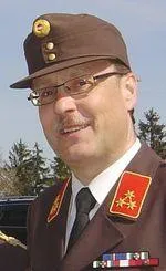 Ein Mann in einem Militäruniform mit einem braunen Hut, Brille, einem roten Abzeichen und mehreren Medaillen steht draußen. Der Hintergrund ist unscharf mit Bäumen.