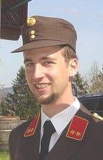 Ein lächelnder Mann in einer Militäruniform mit roten Sternen an den Schultern und einem braunen Hut steht sonnig draußen.