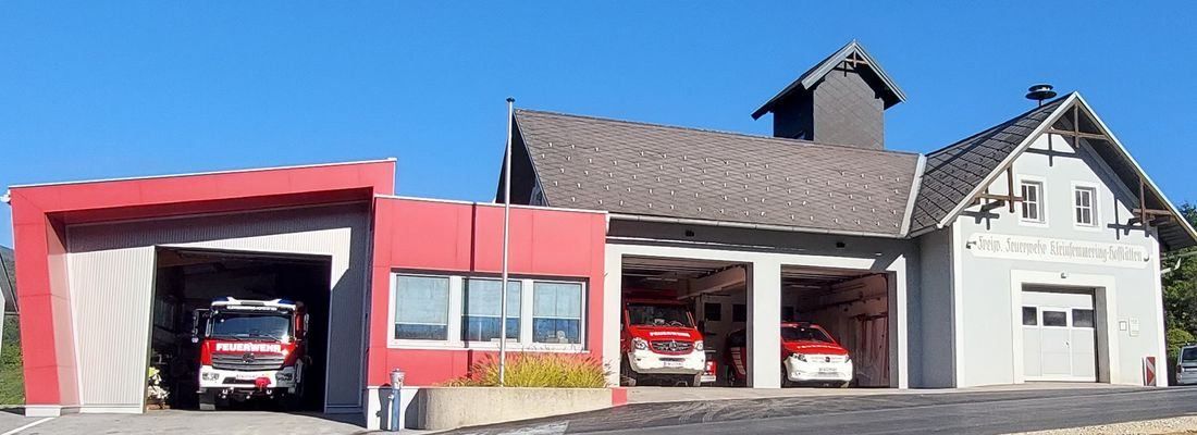 Eine Feuerwehrstation mit einem roten Gebäude, grauem Dach und drei Feuerwehrfahrzeugen, die im Inneren geparkt sind. Die Fahrzeuge sind rot mit weißen Markierungen, und ein Fahnenmast ist vorhanden.