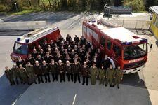 Eine Gruppe Feuerwehrleute in grünen Uniformen steht vor zwei roten Feuerwehrfahrzeugen auf einem Parkplatz. Bäume und ein Zaun sind im Hintergrund.