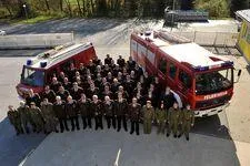 Eine Gruppe Feuerwehrleute in grünen Uniformen steht vor zwei roten Feuerwehrfahrzeugen auf einem Parkplatz. Bäume und ein Zaun sind im Hintergrund.