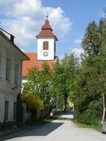 Eine Kirche mit rotem Dach und Turm steht in der Mitte einer Straße, mit Bäumen und einem Gebäude auf beiden Seiten.