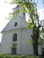Eine weiße Kirche mit einem Kirchturm und einem runden Fenster über dem Haupteingang. Ein Baum mit üppigen grünen Blättern steht davor.