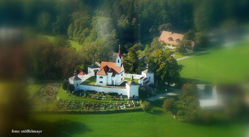 Eine weiße Kirche mit einem roten Dach ist von einem Friedhof umgeben. Die Kirche ist von Bäumen und grünen Feldern umgeben. Ein Haus in der Ferne ist ebenfalls sichtbar.
