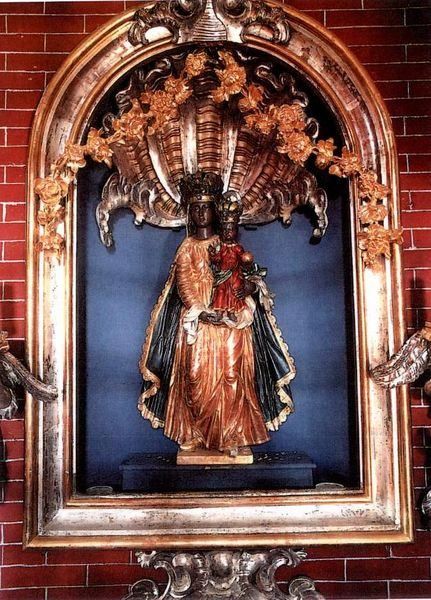 Eine Statue einer Frau, die ein Kind hält, steht in einem kunstvollen Rahmen an einer roten Ziegelmauer, geschmückt mit komplizierten goldenen Mustern.