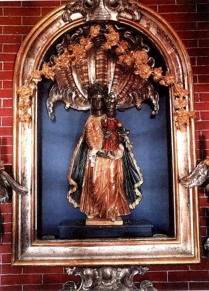 Eine Statue einer Frau, die ein Kind hält, steht in einem kunstvollen Rahmen an einer roten Ziegelmauer, geschmückt mit komplizierten goldenen Mustern.