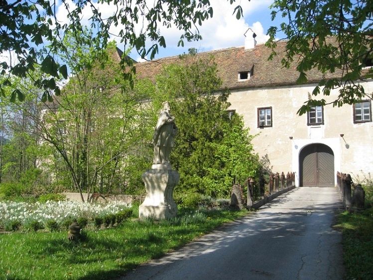 Ein altes Gebäude mit einer Statue davor, umgeben von Grün, und ein Weg führt zu einem Holztor.
