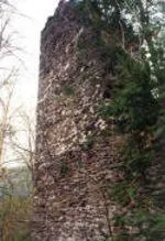 Ein alter, verwitterter Steinturm steht inmitten eines dichten Waldes. Die Oberfläche des Turms ist mit Moos und Efeu bedeckt, und mehrere kleine Löcher sind sichtbar.