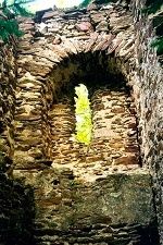 Eine verwitterte Steinmauer zeigt ein bogenförmiges Fenster. Das Licht scheint durch. Die Mauer besteht aus unregelmäßigen Steinen, und Moos wächst an den Kanten.